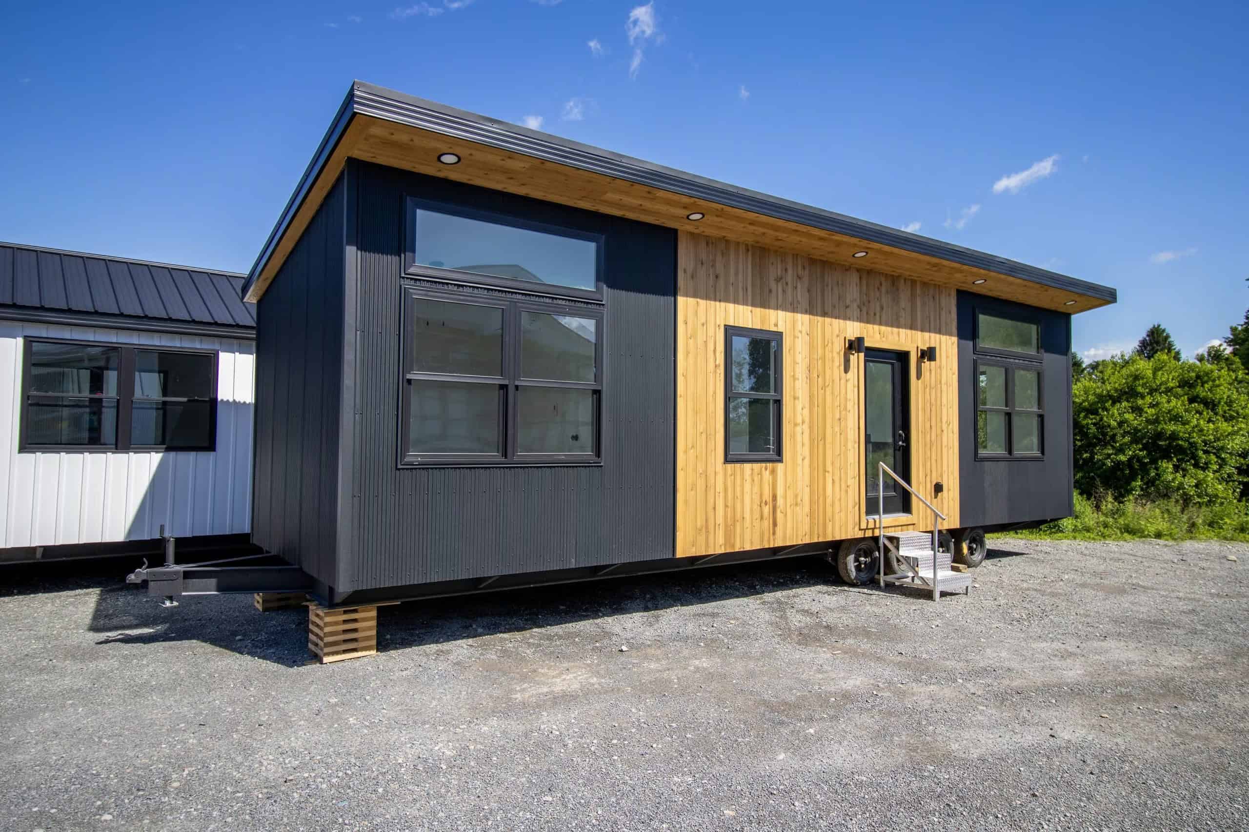 Modern Nova tiny house on a gravel lot: black corrugated metal, cedar around a full-glass entry door, black-framed windows, puck lights in the wood soffit
