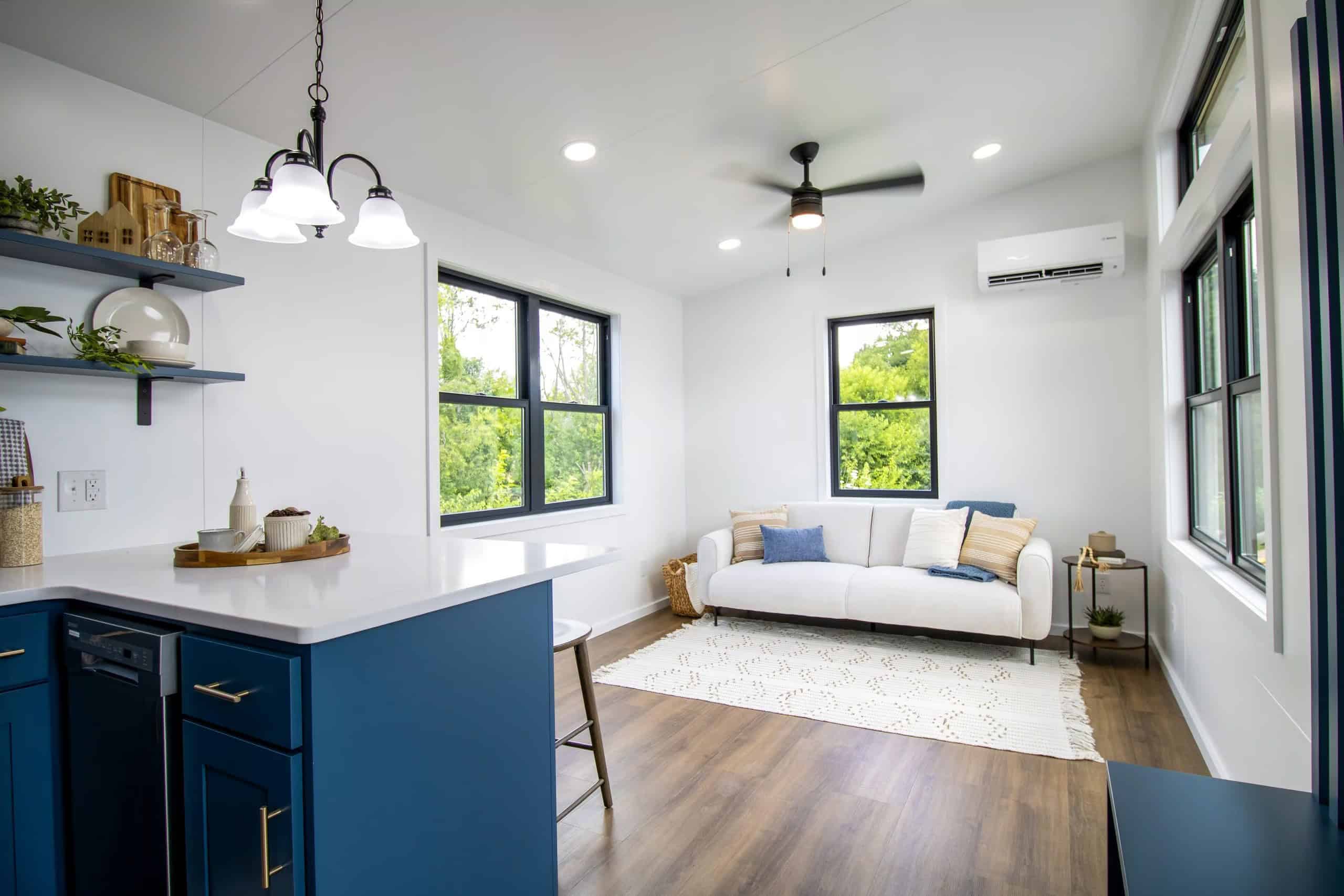 Open living end of the Modern Nova: white sofa under a black-framed window, navy kitchen peninsula with white quartz, black chandelier, Bosch head on the far wall