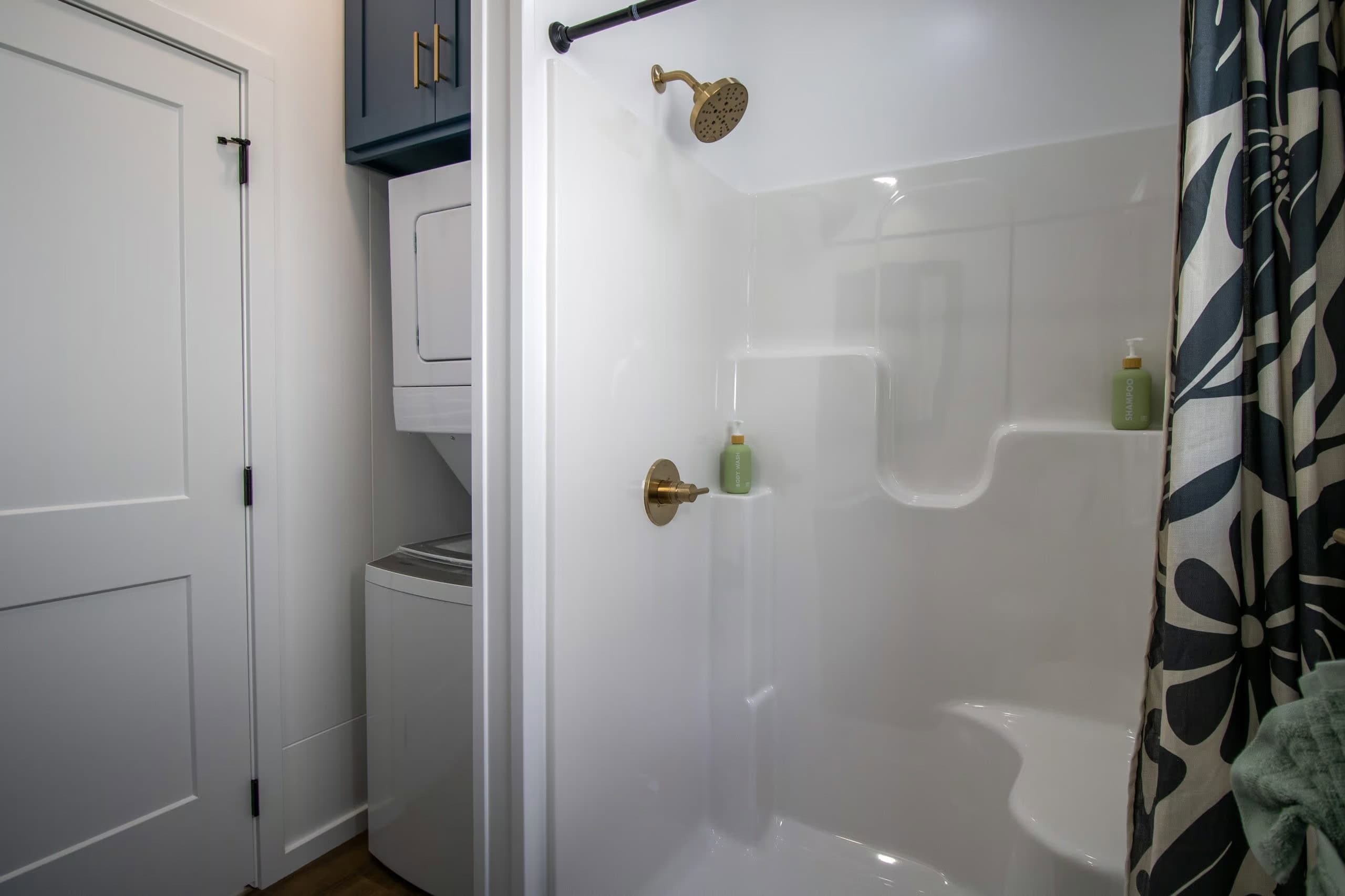 Bathroom: white 4-by-3 fiberglass shower with gold fixtures and a molded seat, stacked white washer and dryer under a navy cabinet