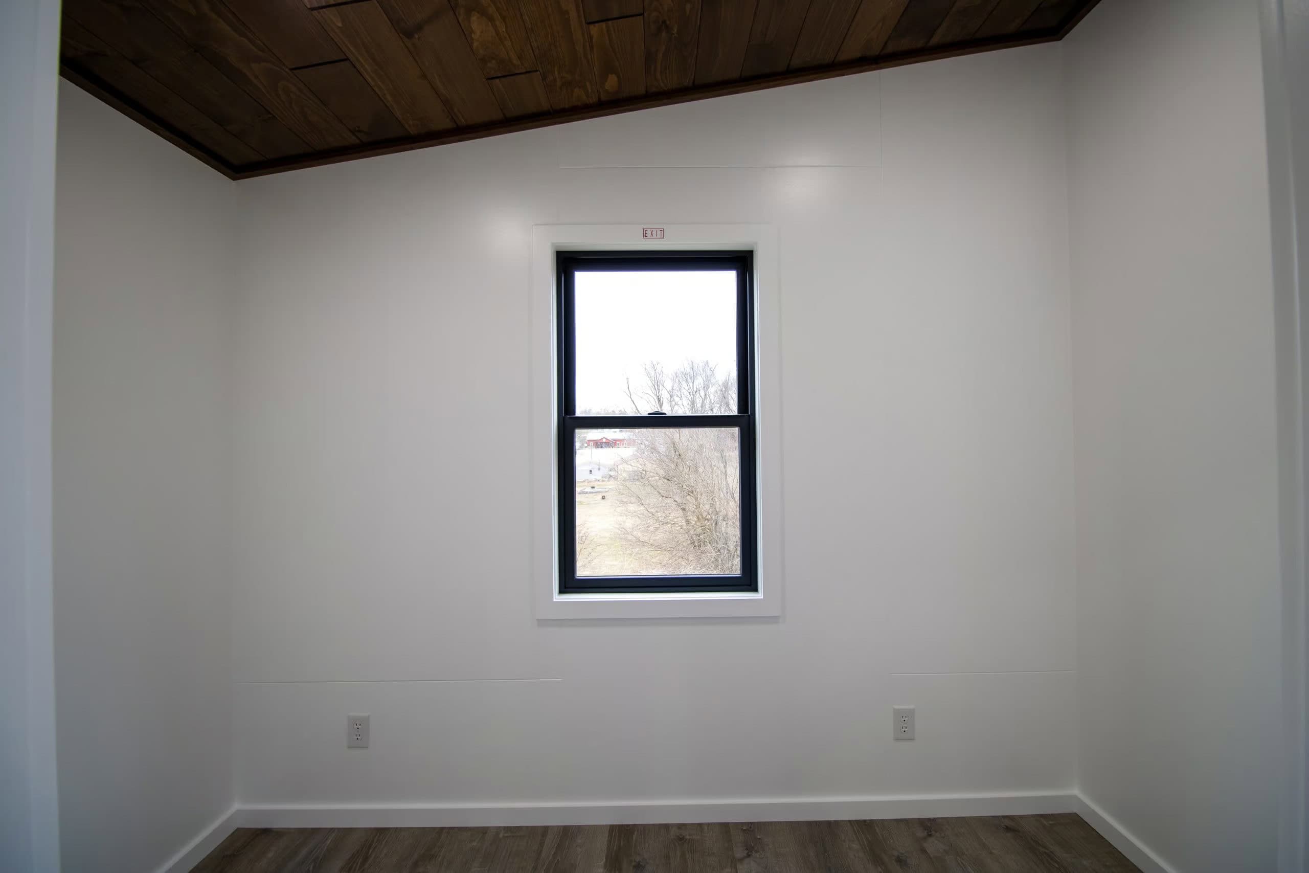 Empty main-floor bedroom: white walls, dark wood ceiling, gray wood-look floor, one black-framed window centered on the wall