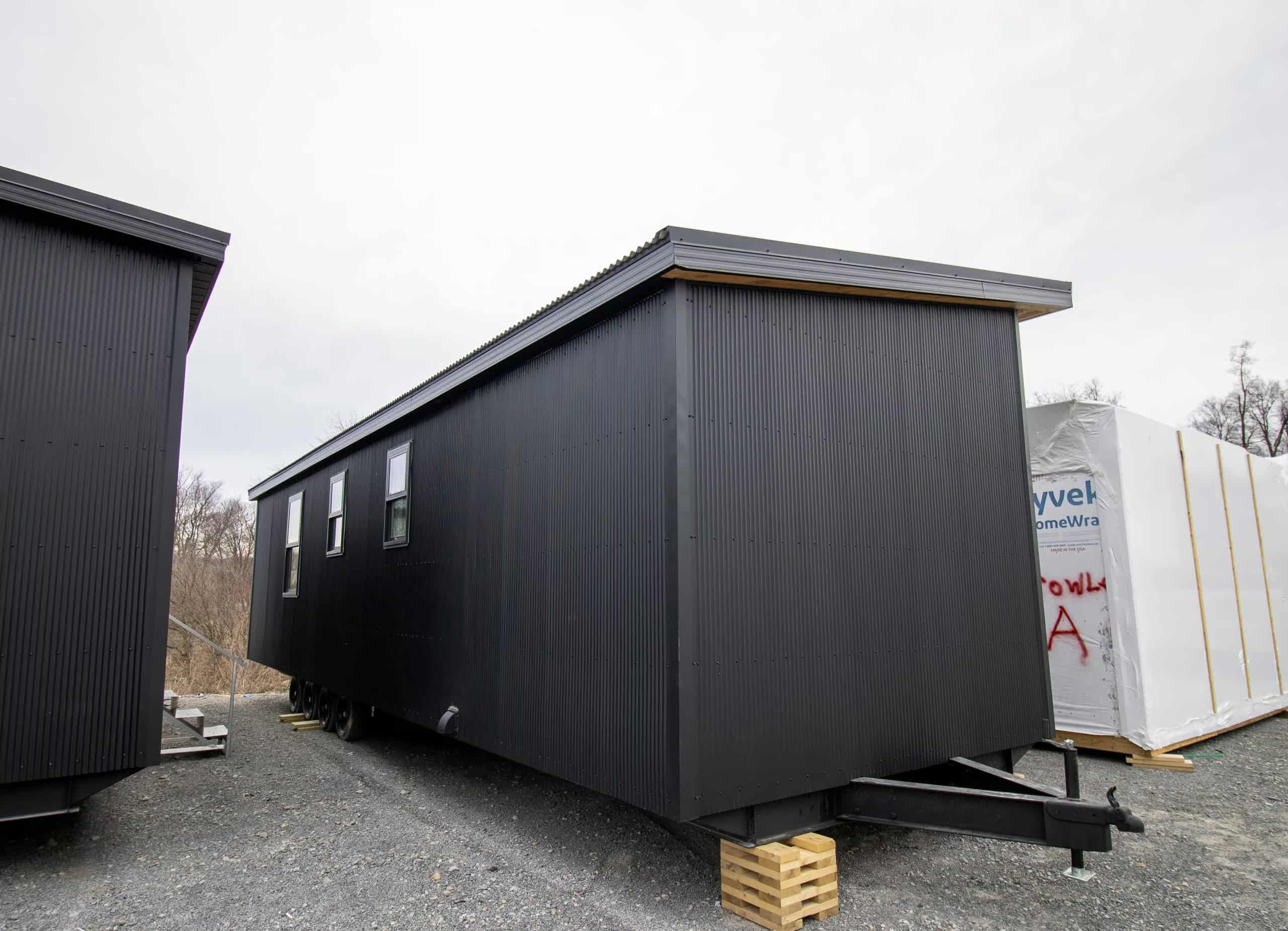 Long black corrugated-metal side of Modern Twilight on a steel trailer, hitch at the near end, three black-framed windows, wood cribbing on gravel