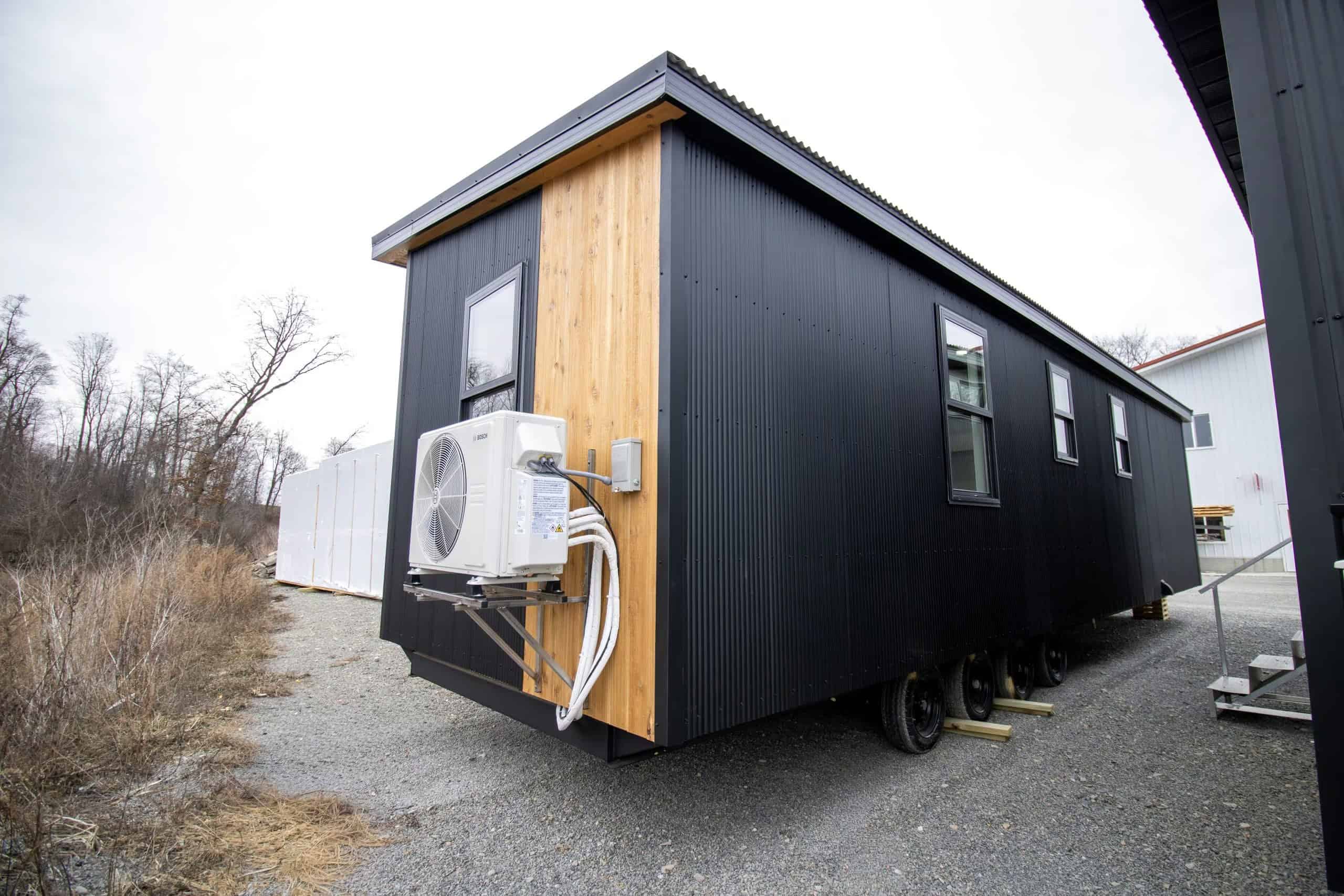 Short end of Modern Twilight: cedar accent panel, black corrugated metal, white Bosch mini-split on a bracket, multi-axle trailer on gravel