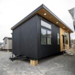 Modern Twilight tiny house on a gravel lot: black corrugated metal, cedar around the glass entry, detachable hitch, wood cribbing under the trailer