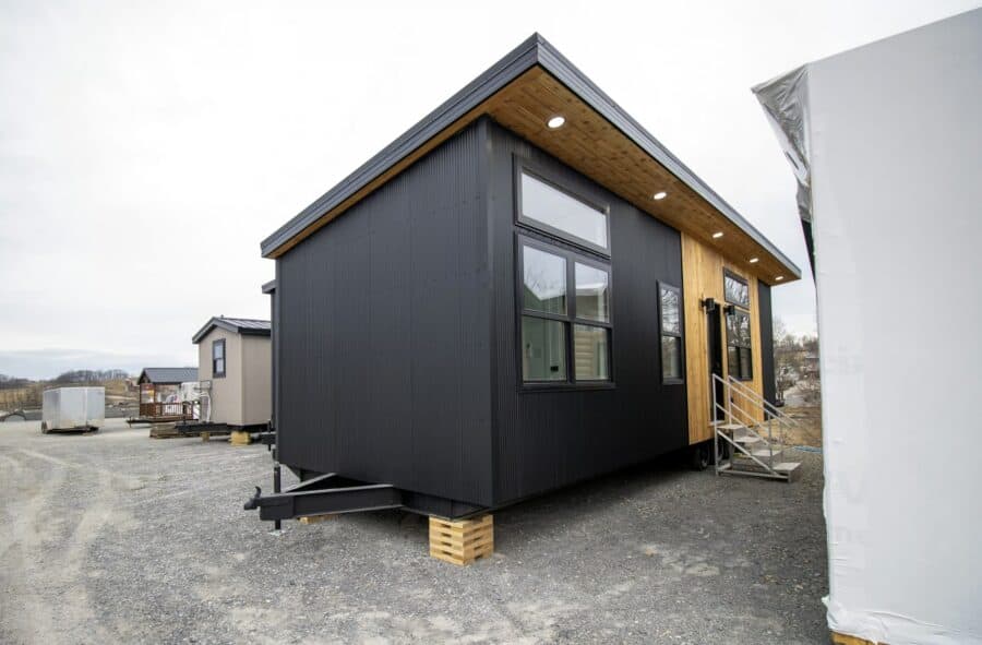 Modern Twilight tiny house on a gravel lot: black corrugated metal, cedar around the glass entry, detachable hitch, wood cribbing under the trailer