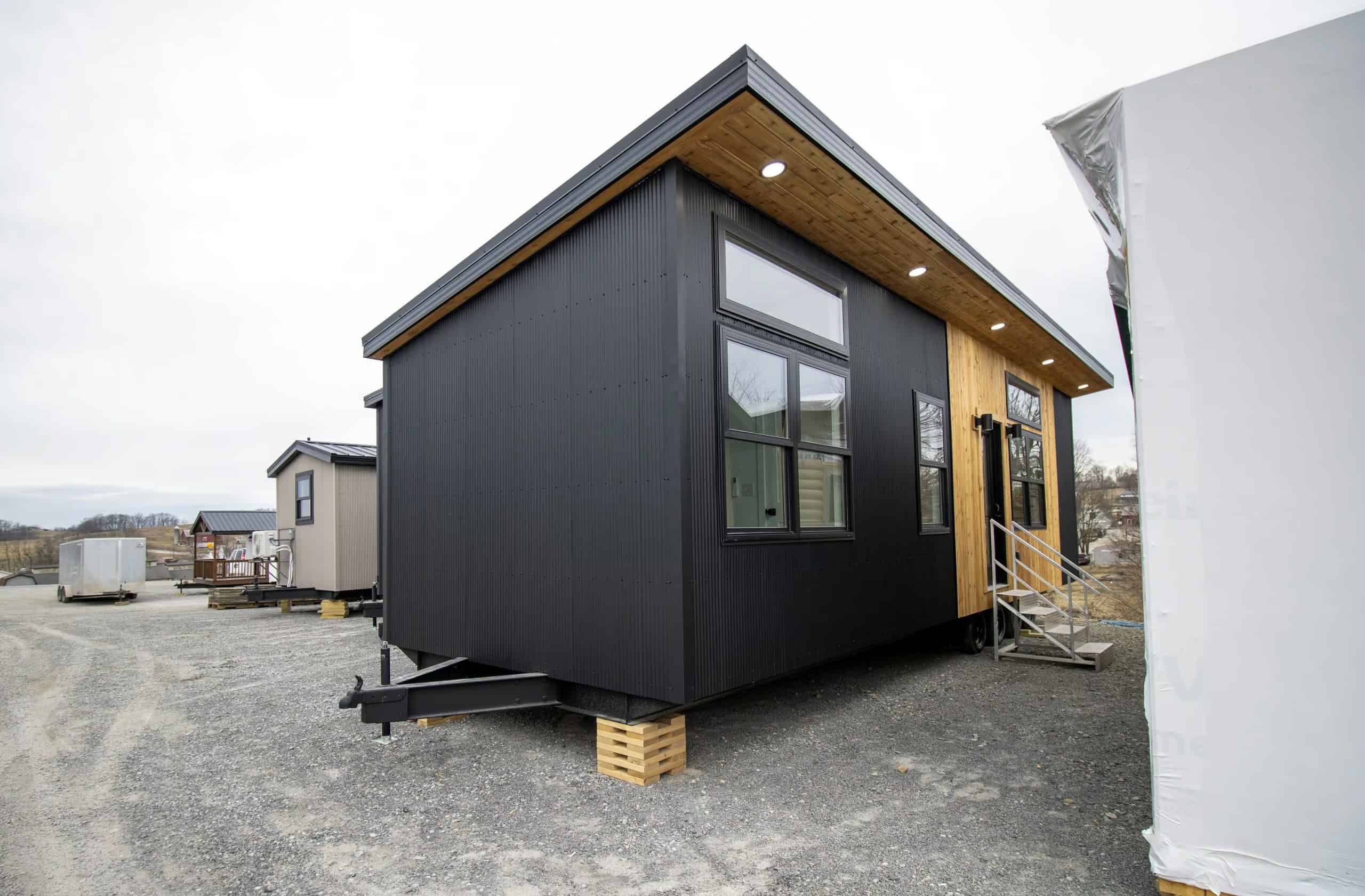 Modern Twilight tiny house on a gravel lot: black corrugated metal, cedar around the glass entry, detachable hitch, wood cribbing under the trailer