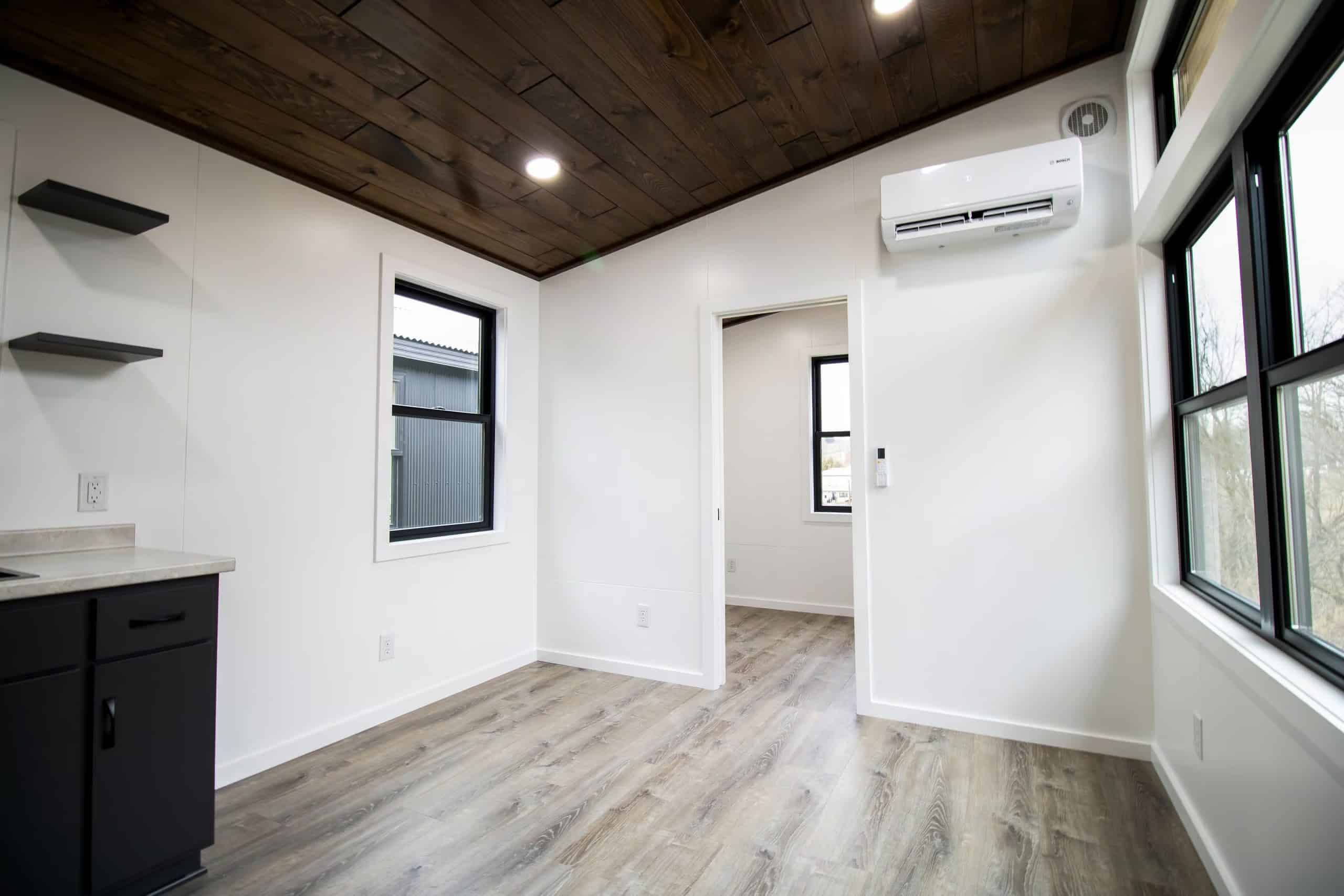 Painted-white interior: dark wood ceiling, wood-look plank floor, black-framed windows, dark cabinets with a light counter, white Bosch head by a doorway
