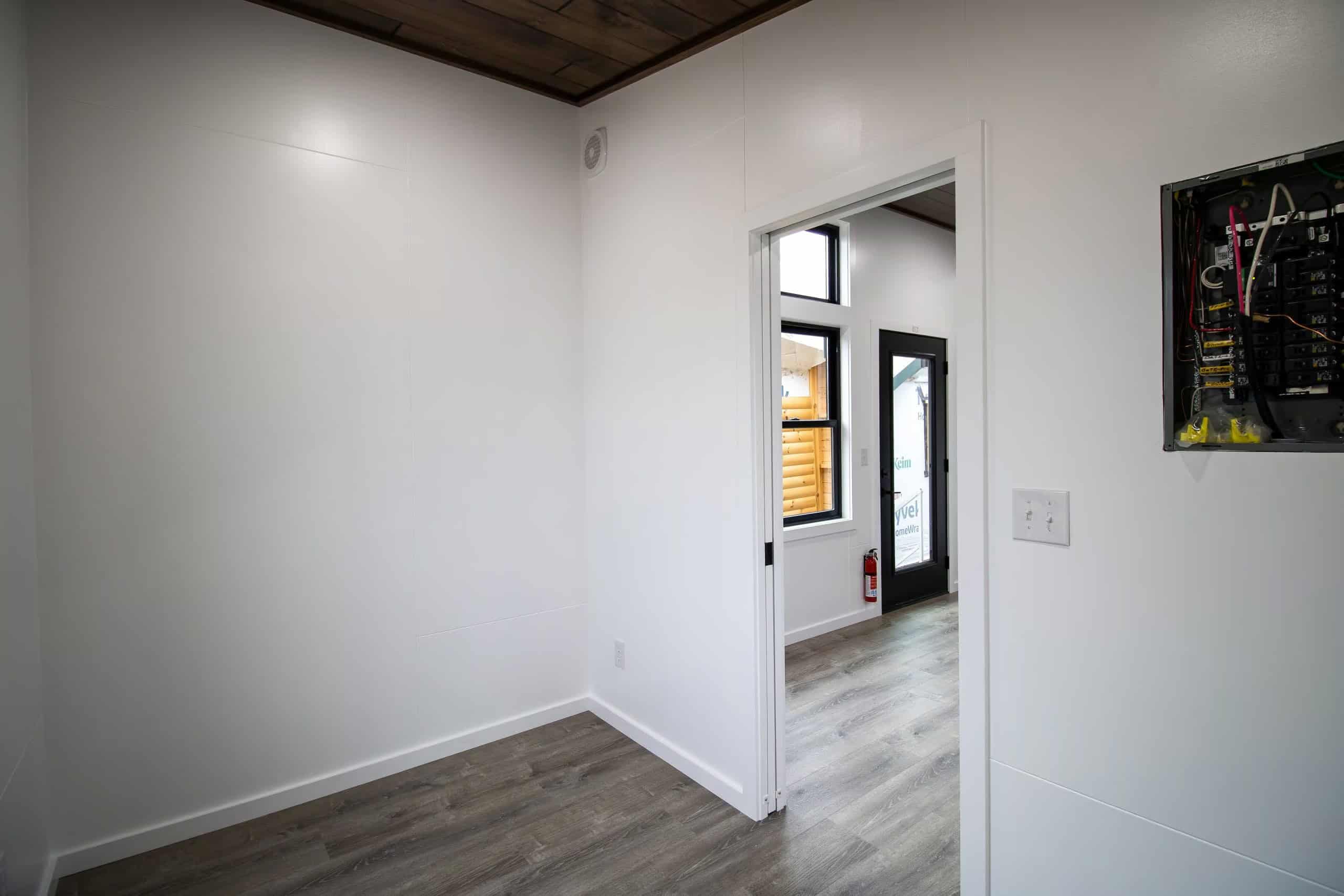 Second main-floor room looking through a doorway to the glass entry, white walls, dark wood ceiling, fire extinguisher on the far wall