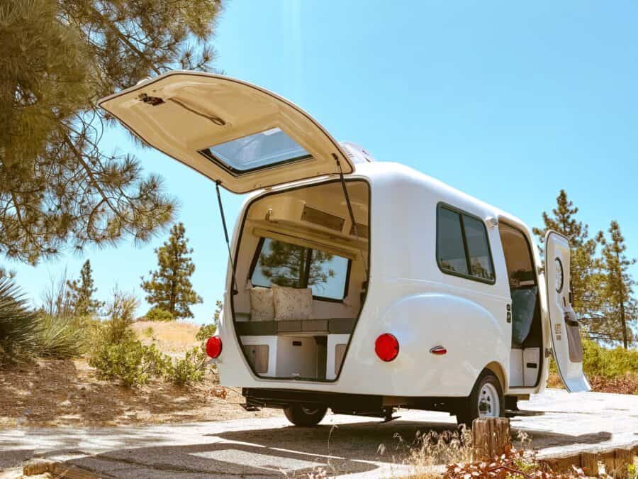 Cream-white Happier Camper HC1 on a gravel path among pines, rear hatch open on gas struts, grey cushions and cream pillows inside, side door open, red round taillights
