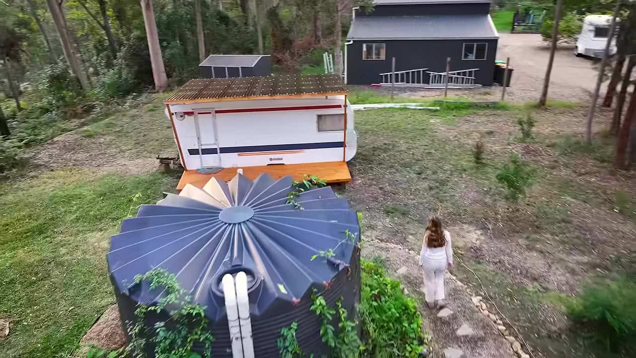 White renovated caravan with a red stripe and a small timber porch beside a large water tank in New South Wales bush