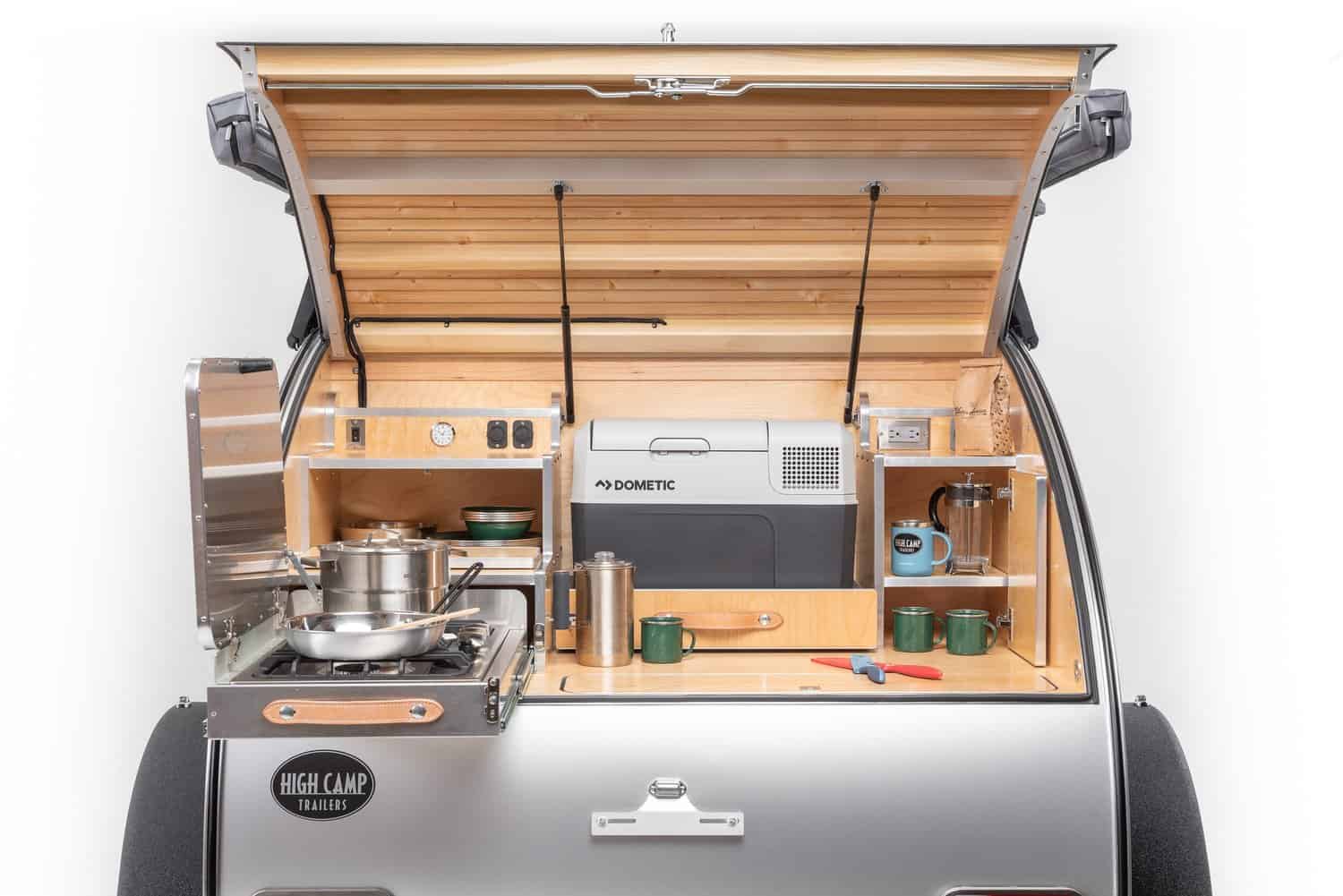 High Camp teardrop rear galley open under the hatch, with wood cabinets, a portable fridge, cookware, and the slide-out stove