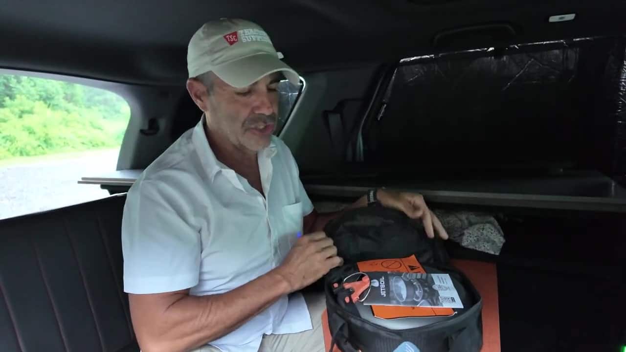 Aaron opening a round zippered JetBoil cook-system case on his lap inside the Honda Pilot camper, with bed platform and blackout covers behind him