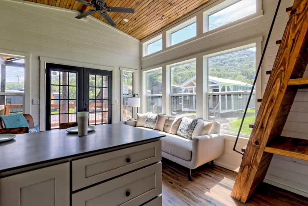 Vaulted wood-ceiling living room with a grey sofa, kitchen island, French doors, and windows looking at neighboring tiny homes