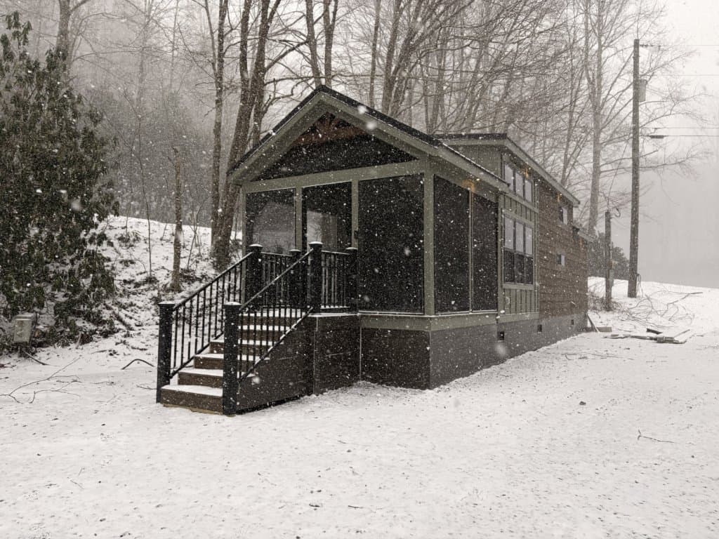 Gabled brown-sided tiny home with a sage porch and dark steps in falling snow