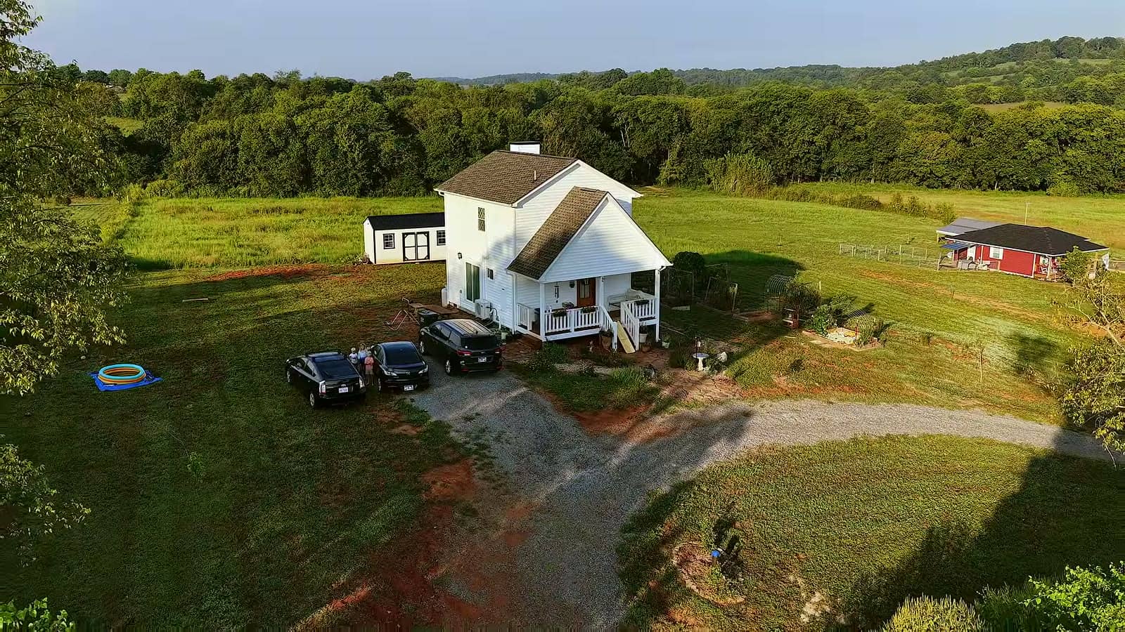 Aerial view of a white two-story tiny farmhouse with front porch, gravel driveway, parked cars, and a small outbuilding in green Tennessee fields