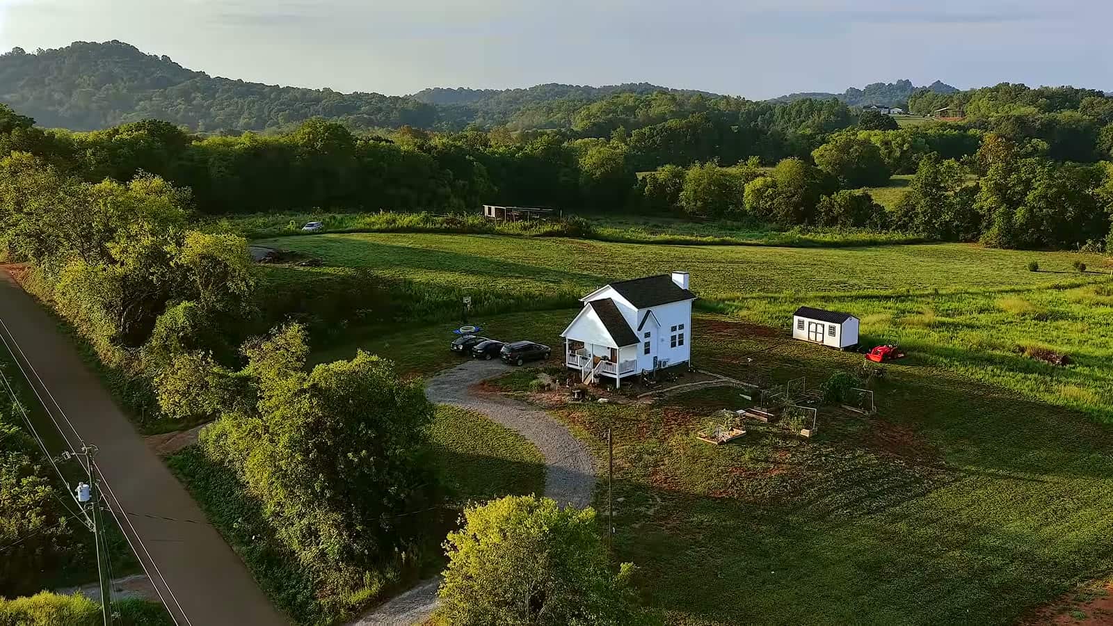 Small white farmhouse in a wide green valley surrounded by trees and hills at sunset