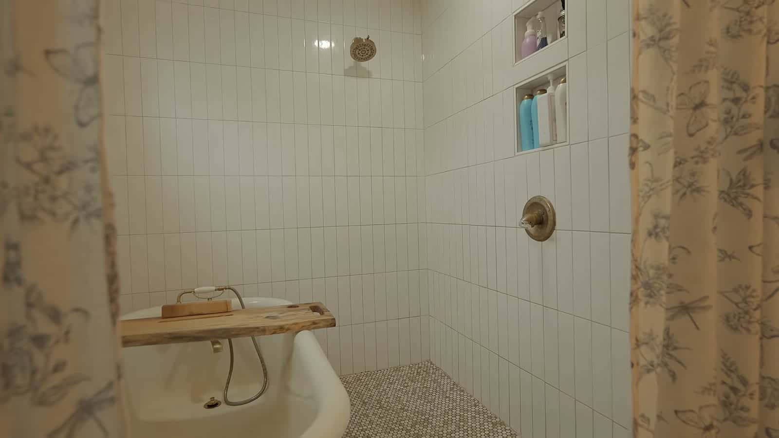 Clawfoot tub inside a white-tiled walk-in shower with penny tile floor, wall niche, and floral shower curtain