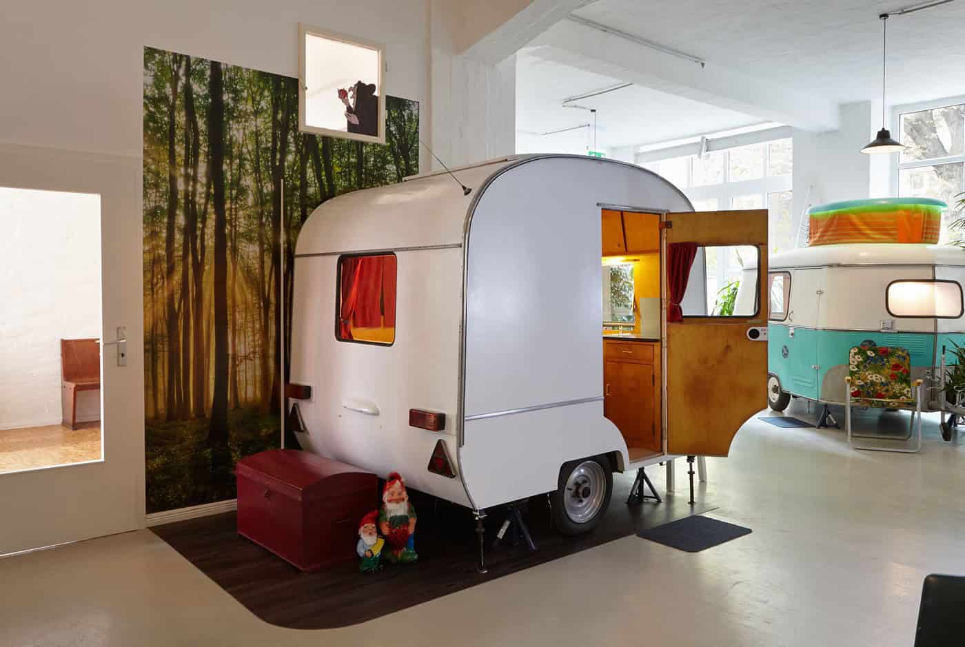White vintage caravan indoors with the door open, forest mural behind — not a named Hall 1 unit