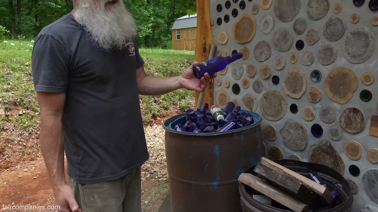 Bearded man holding a blue glass bottle beside a barrel full of blue bottles, next to a cordwood and bottle wall