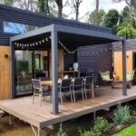 Two dark corrugated-metal tiny-house pods connected by a raised timber deck with outdoor dining under a pergola, on a lawn against Blue Mountains forest