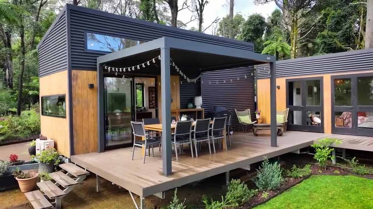 Two dark corrugated-metal tiny-house pods connected by a raised timber deck with outdoor dining under a pergola, on a lawn against Blue Mountains forest