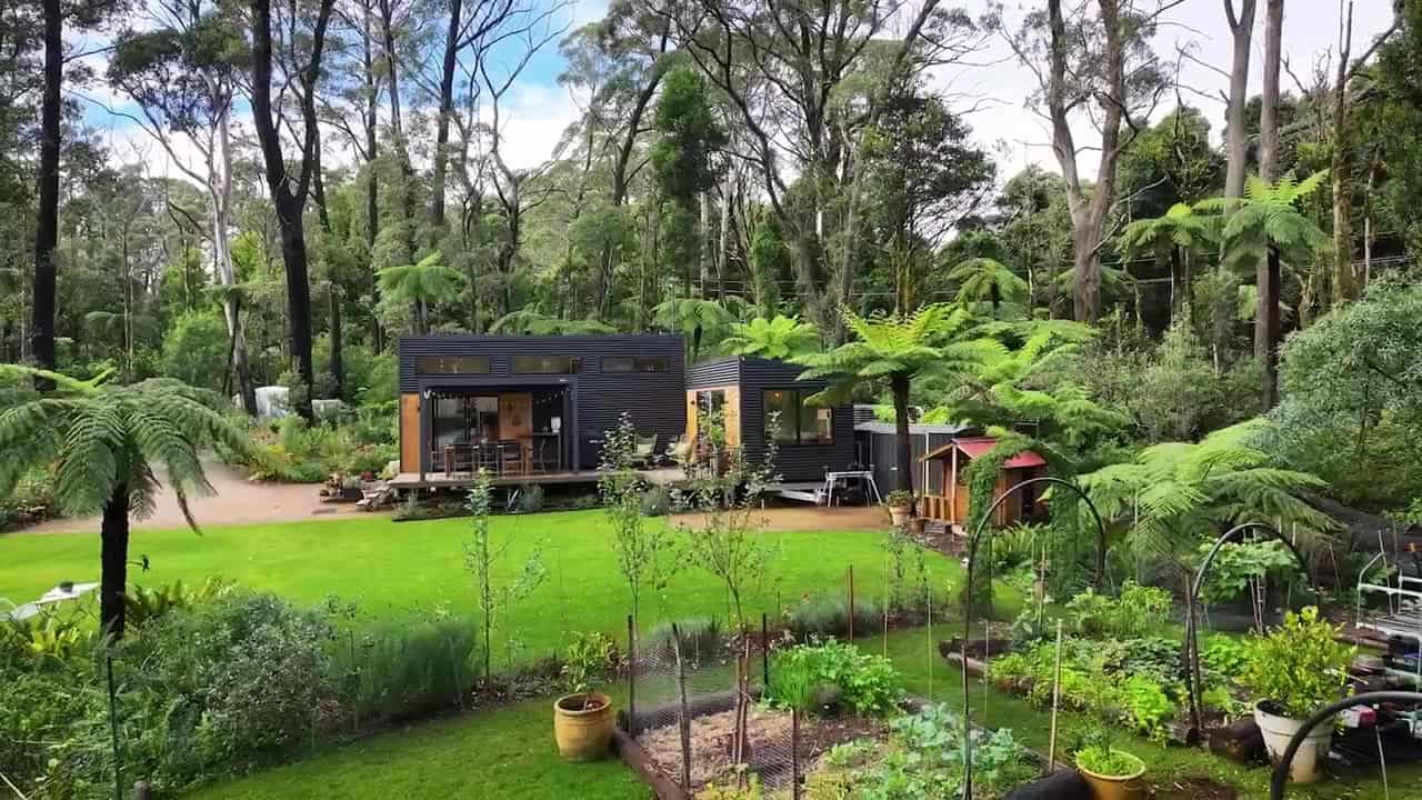 Raised vegetable beds in front of two dark metal tiny-house pods and a timber deck on a lawn, with forest behind and trailer wheels under the right-hand pod