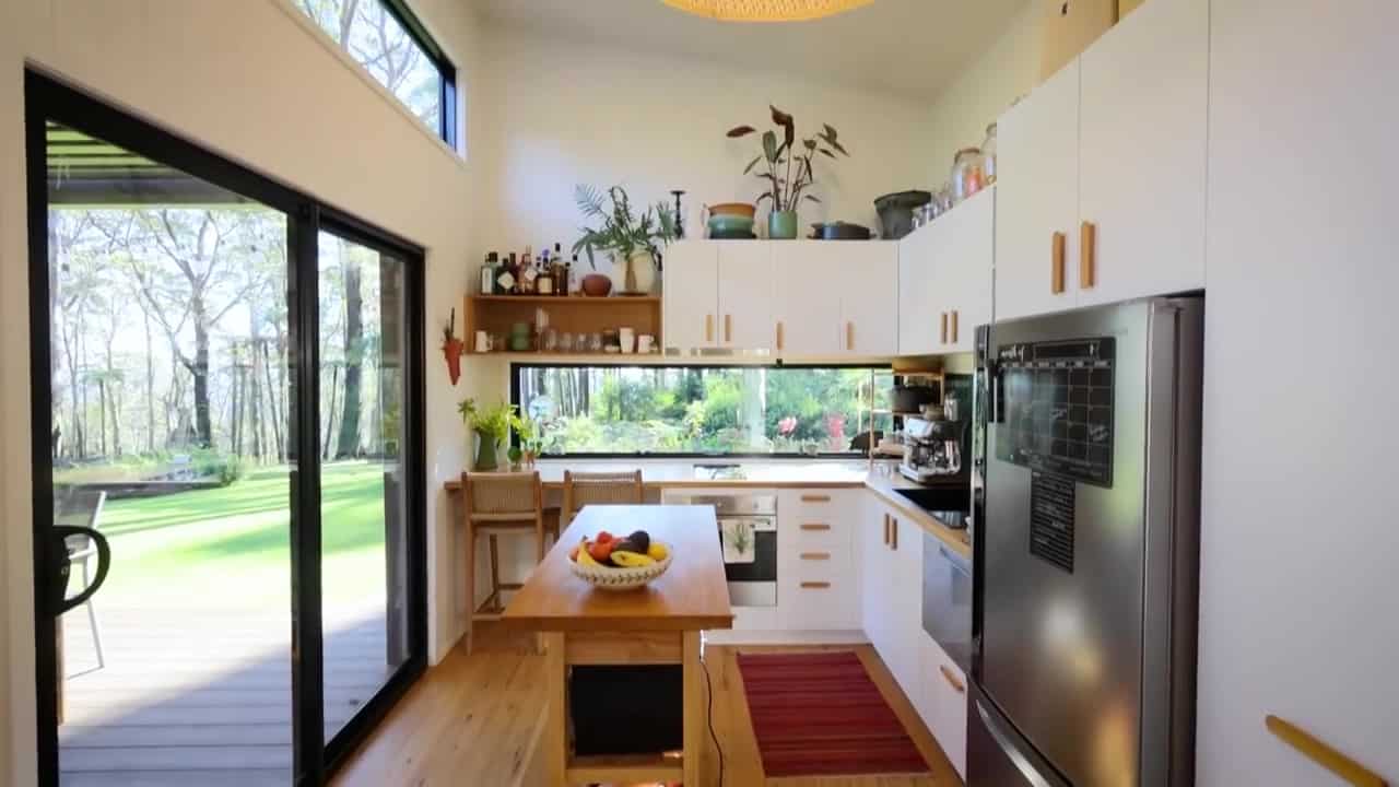 L-shaped tiny-house kitchen with white cabinets, wood counters, green tile, a long window, island, breakfast bar with rattan stools, and a full-size fridge