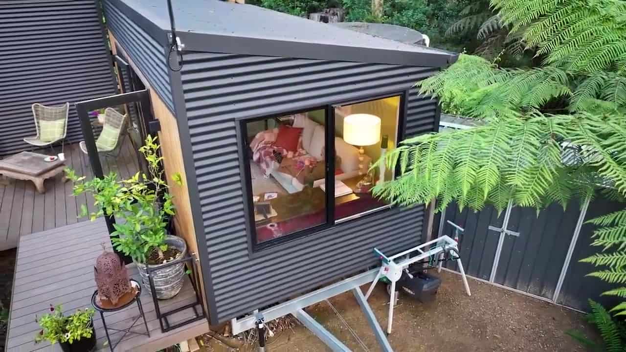 Dark corrugated-metal living pod on a trailer chassis with stabilizer jacks, a large window into a sofa, and a timber deck with green chairs