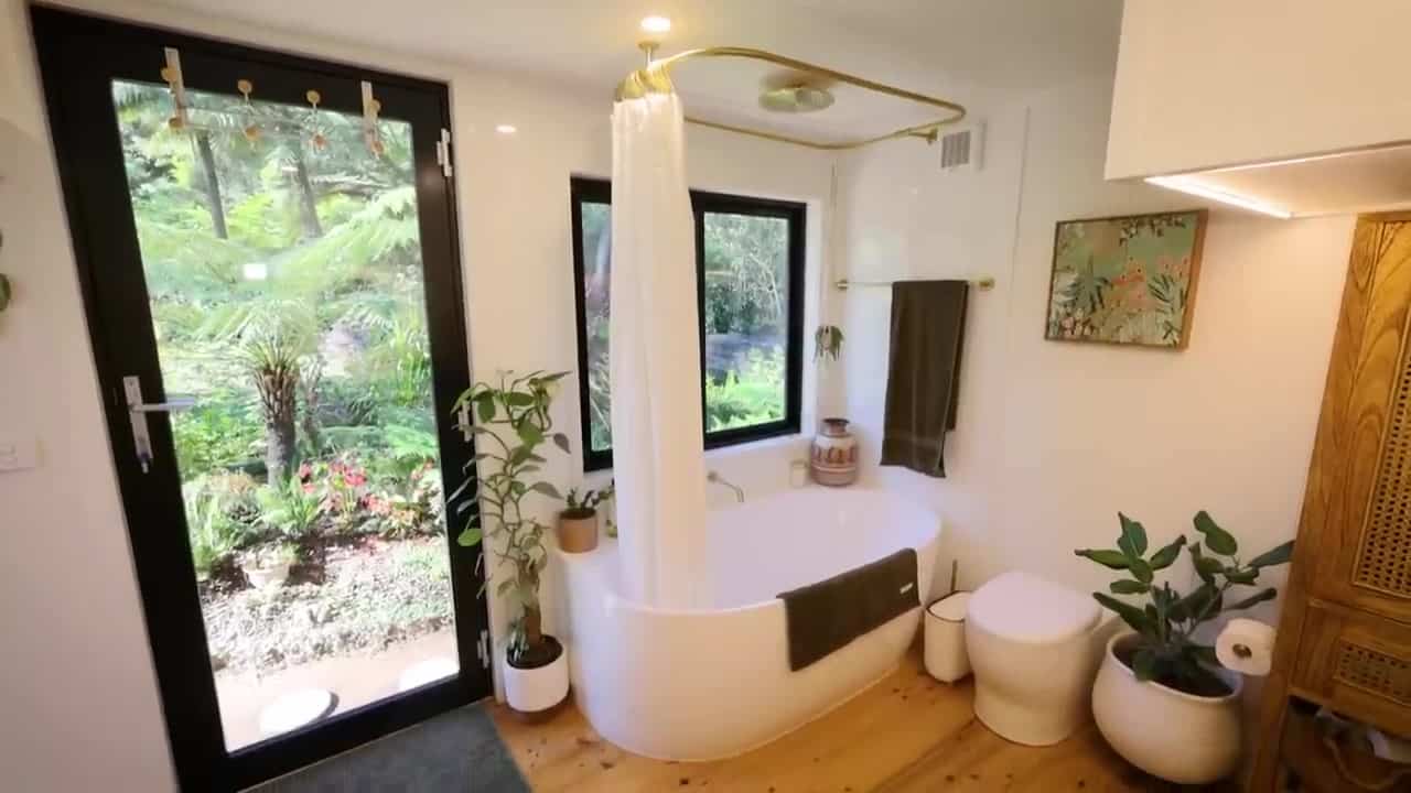 White freestanding bathtub under a window looking into ferns, with a glass garden door, brass shower rail, and teak storage in a bright tiny-house bathroom