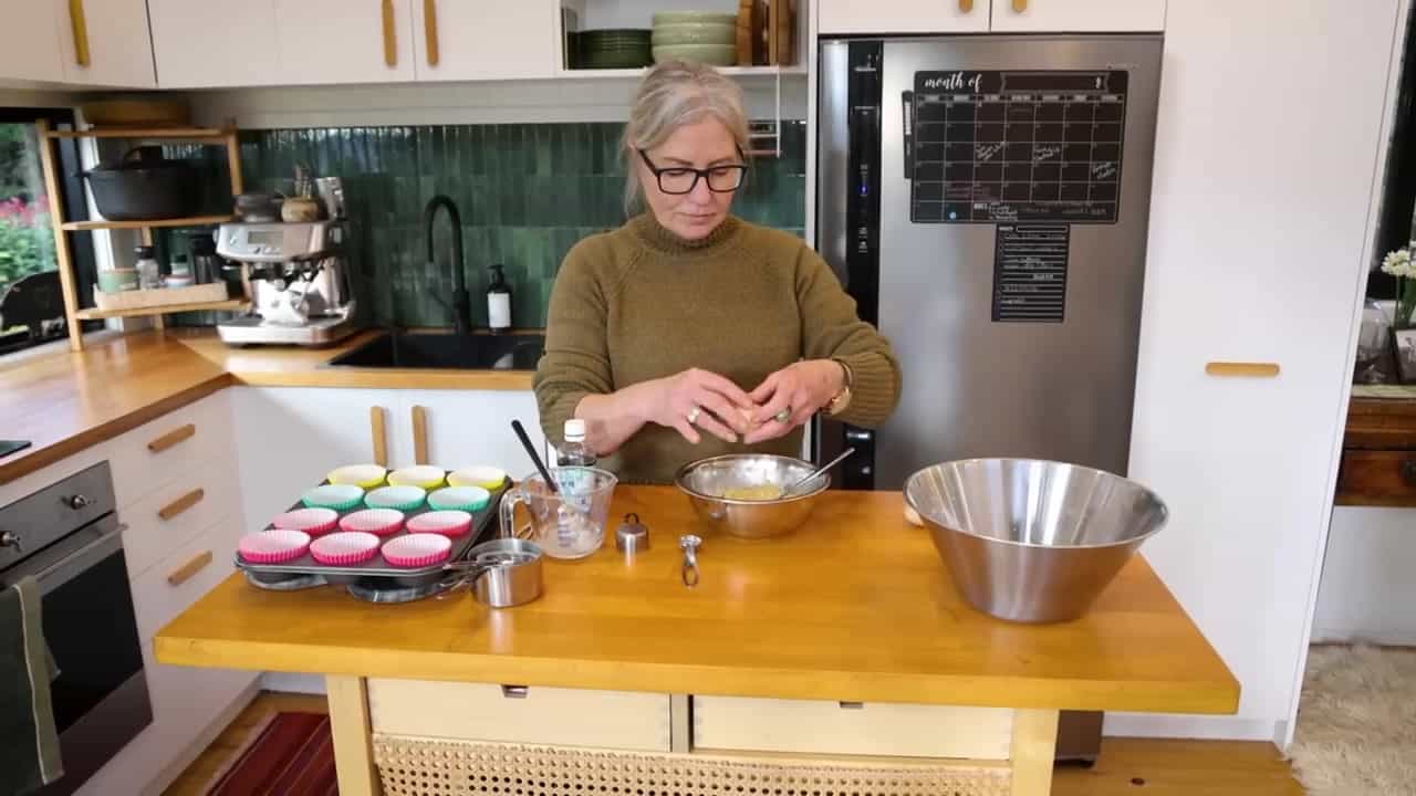 Joanne at a wood island cracking an egg into a bowl, with a muffin tin, green tile backsplash, espresso machine, and full-size fridge behind her