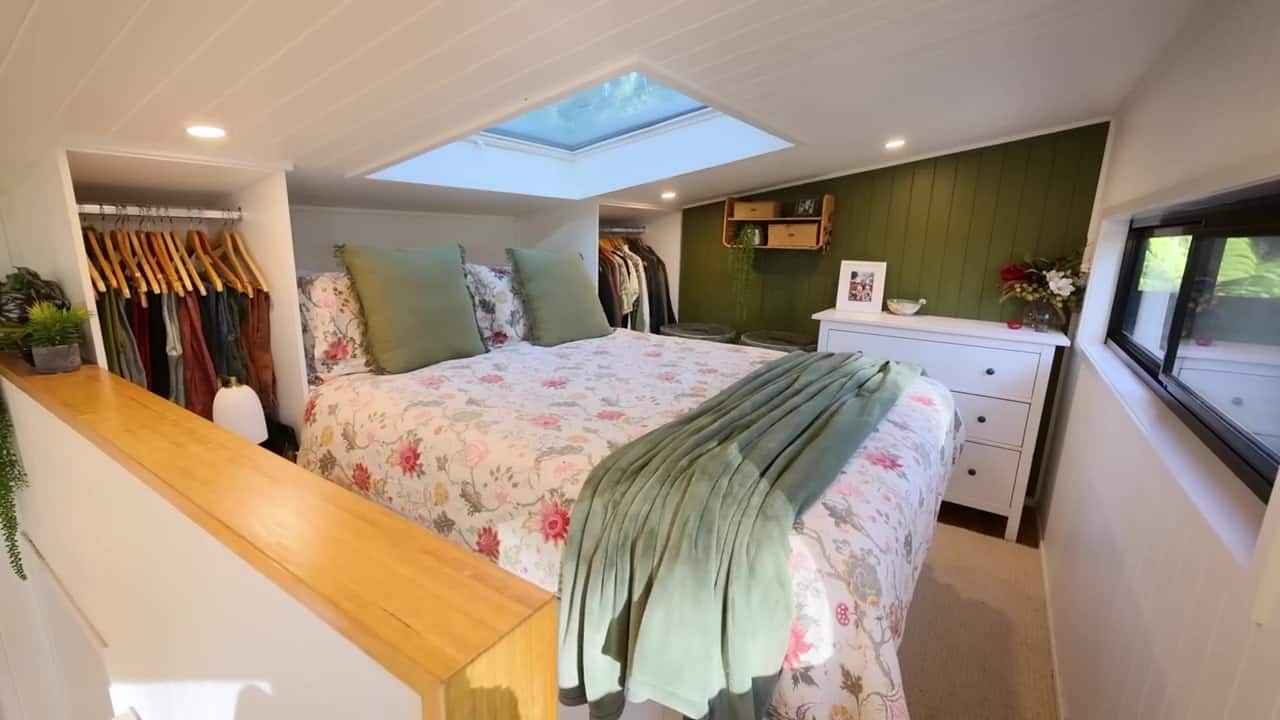 Standing-height tiny-house loft with a floral bed, skylight over the trees, hanging storage on both sides, and a sage-green feature wall