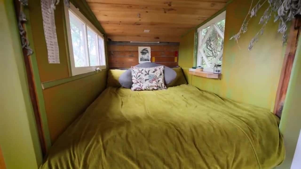 Inside the popped-out bed nook with olive quilt, side windows, and a small shelf toward the trees