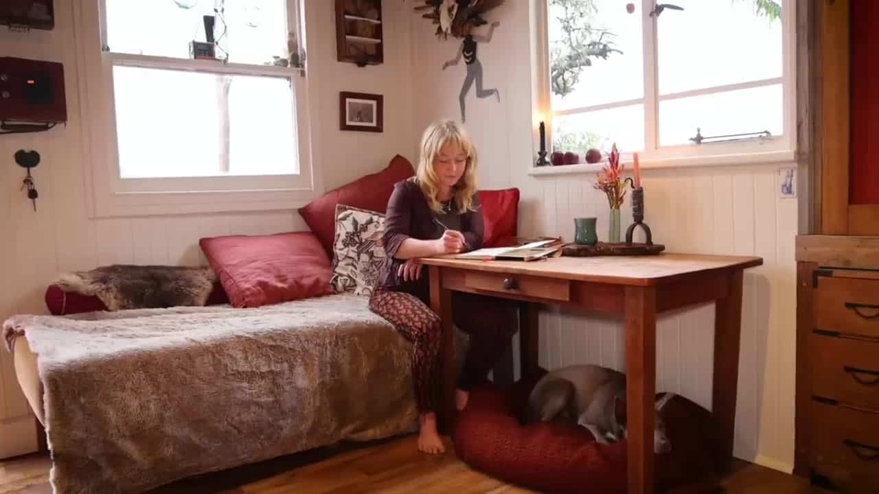 Kayla drawing at a small wooden table, dog Jemba asleep on a red cushion underneath, daybed by the windows