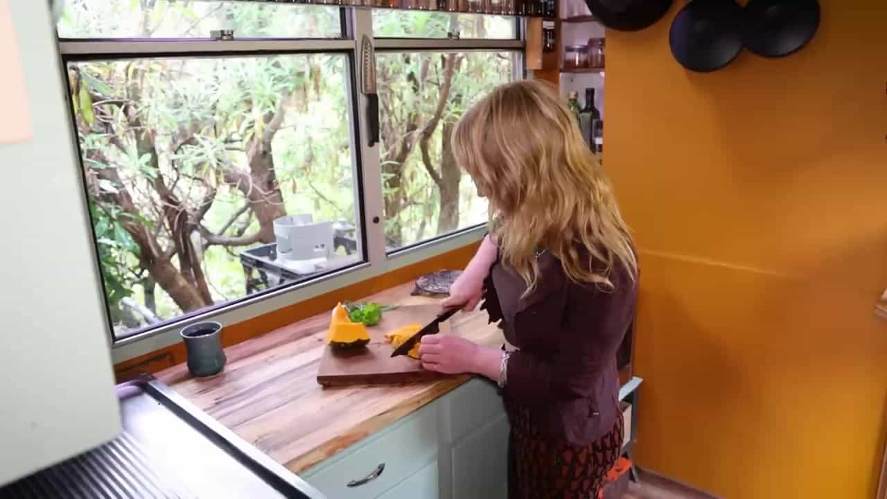 Kayla chopping squash at a wooden counter in a small orange kitchen with a wide window into the trees