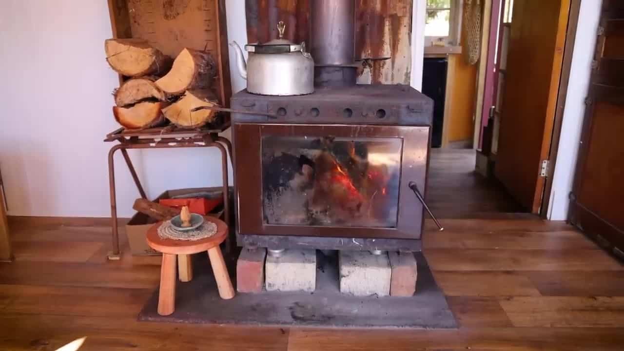 Wood stove with a kettle on top, fire burning, rusty corrugated steel chimney backing salvaged from a bushfire