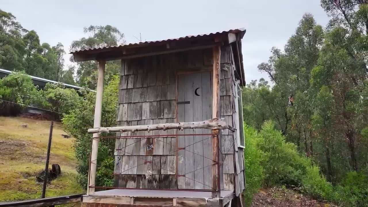 Weathered composting outhouse with hand-split shingles, rusty roof, and a crescent moon cut into the door, Tasmanian bush behind