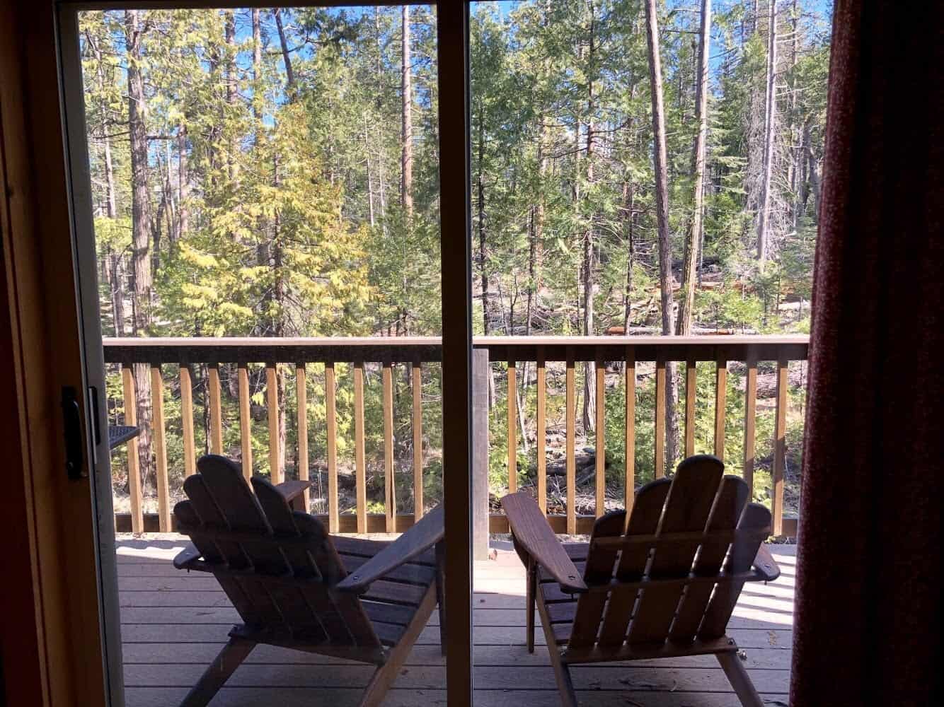 Two empty chairs on a wood deck, pines beyond the glass