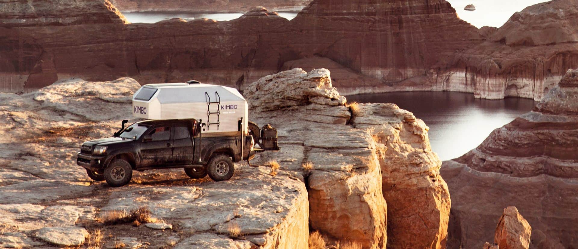 Silver Kimbo 6 hard-side on a dark midsize pickup parked on a rock shelf at golden hour, canyon water behind, KIMBO on the cab-over and a side ladder