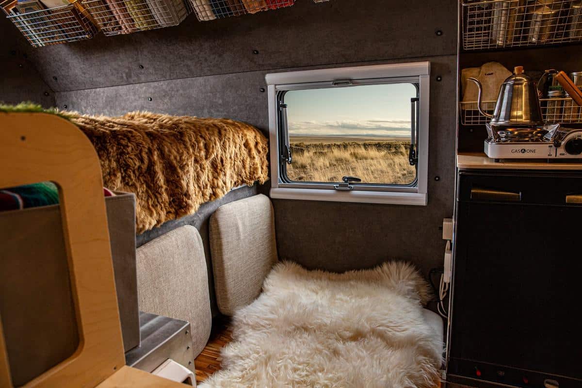 Kimbo 6 cab-over interior: dark riveted walls, white-framed Arctic Tern window on yellow grass, sheepskin on the floor, wire baskets and a kettle on the counter