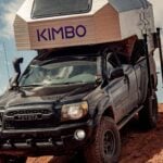 Silver faceted Kimbo 6 hard-side truck-bed camper slid into a black midsize pickup on red dirt, blue KIMBO letters on the cab-over, dark rectangular window above