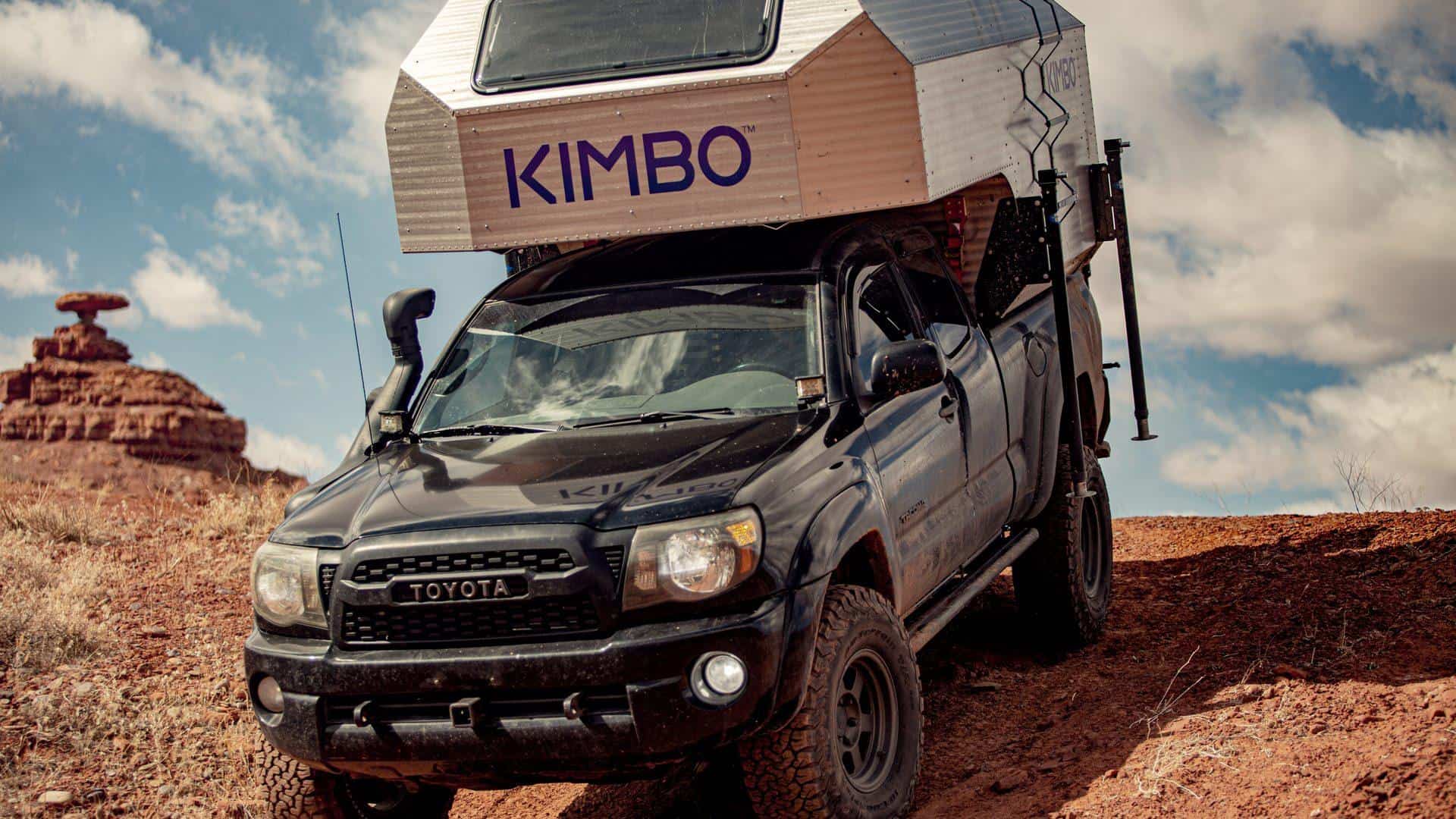 Silver faceted Kimbo 6 hard-side truck-bed camper slid into a black midsize pickup on red dirt, blue KIMBO letters on the cab-over, dark rectangular window above