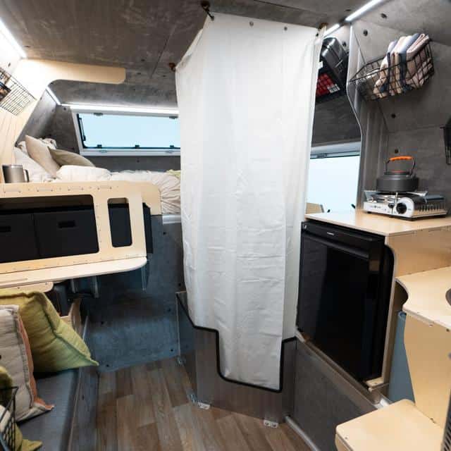 Kimbo 6 interior: loft with white bedding, kitchen module with a small black fridge, LED ceiling strips, white privacy curtain over a riveted aluminum pan
