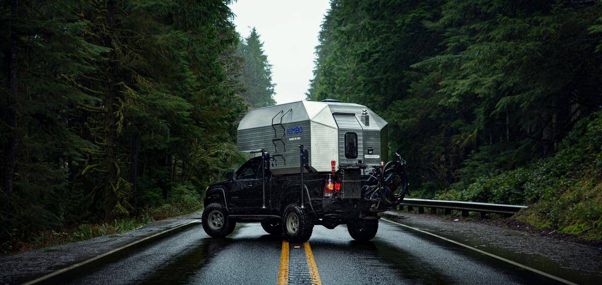 Silver Kimbo 6 on a dark midsize pickup parked across a wet two-lane through evergreens, MADE IN USA on the side, a bike on the rear rack