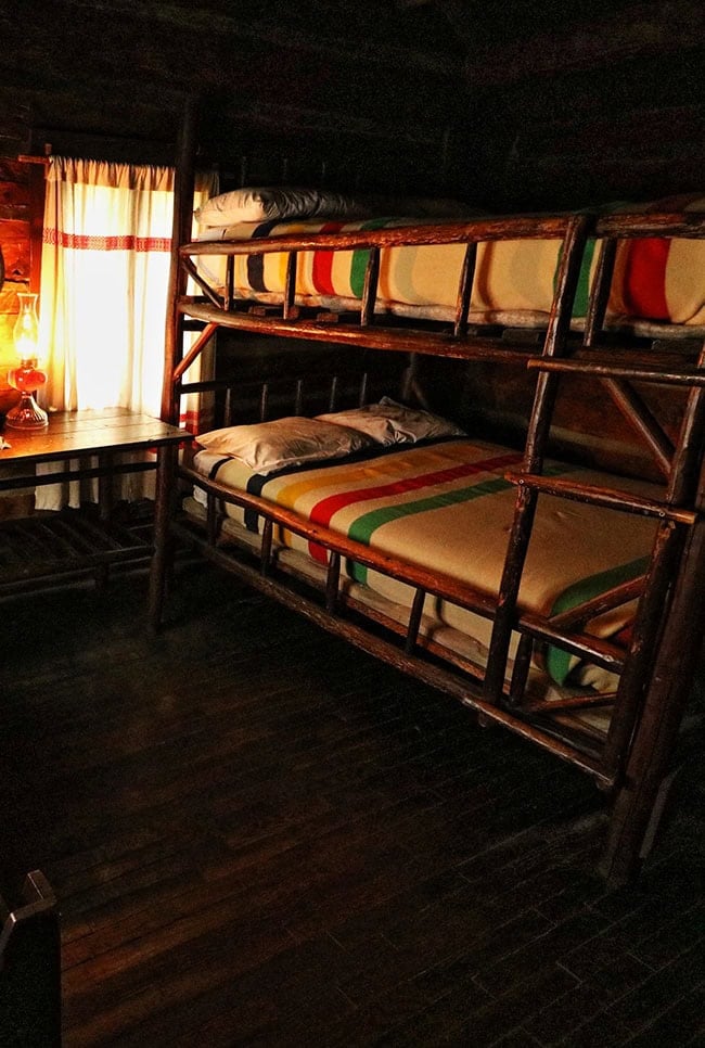 Dark log-cabin bunk interior: rough-hewn log bunks with a branch ladder, white linens and cream Hudson Bay wool blankets with green red and yellow stripes, a lit kerosene lantern on a small table by a curtained window
