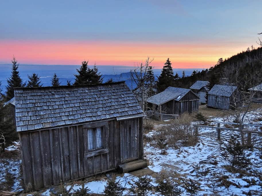 Small shake-roofed cabins on a snowy slope at dusk, an orange-pink horizon over distant ridges, spruce silhouettes, a rail fence at right