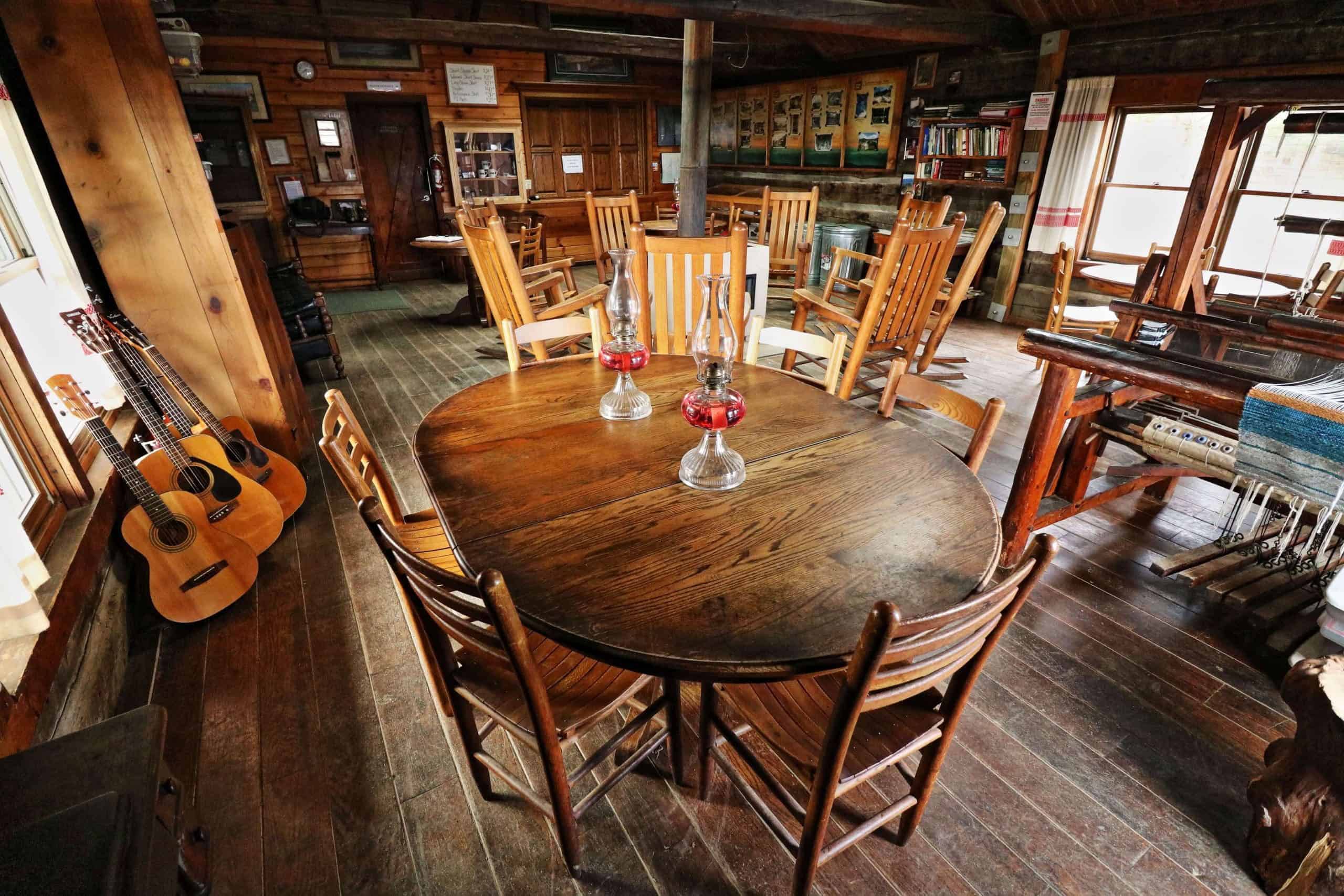 Lodge common room: oval dining table with two red-base lanterns and four slat-back chairs, three guitars by a window, a floor loom with a blue-and-white weaving, rocking chairs, a stove pipe, and a packed bookshelf