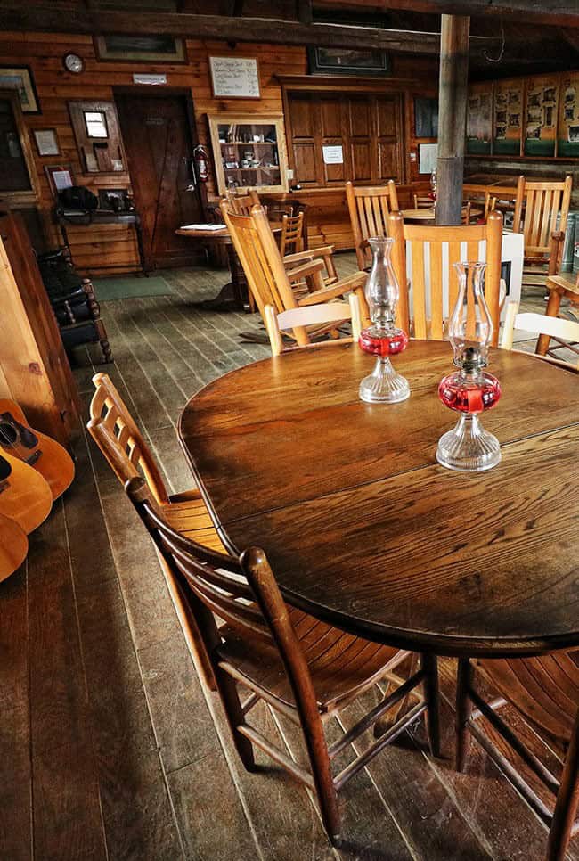 Lodge dining and common room: a round dark-wood table with two red-base kerosene lanterns, slat-back chairs, acoustic guitars leaning at left, a black stove pipe rising through the room, rocking chairs beyond