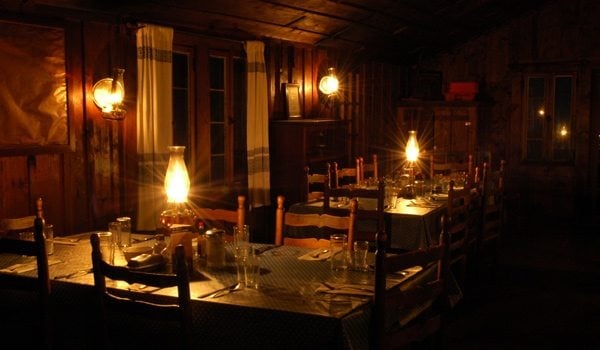 Dining hall at night: two long tables with blue-and-white checkered cloths, glassware and silverware, kerosene lanterns on the tables and mounted on dark wood walls