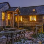 LeConte Lodge at dusk: weathered shake siding and a wood-shingle roof, warm light in the multi-pane windows, a log fence and picnic table in front, snow patches on mossy ground, a wooden walkway to the right