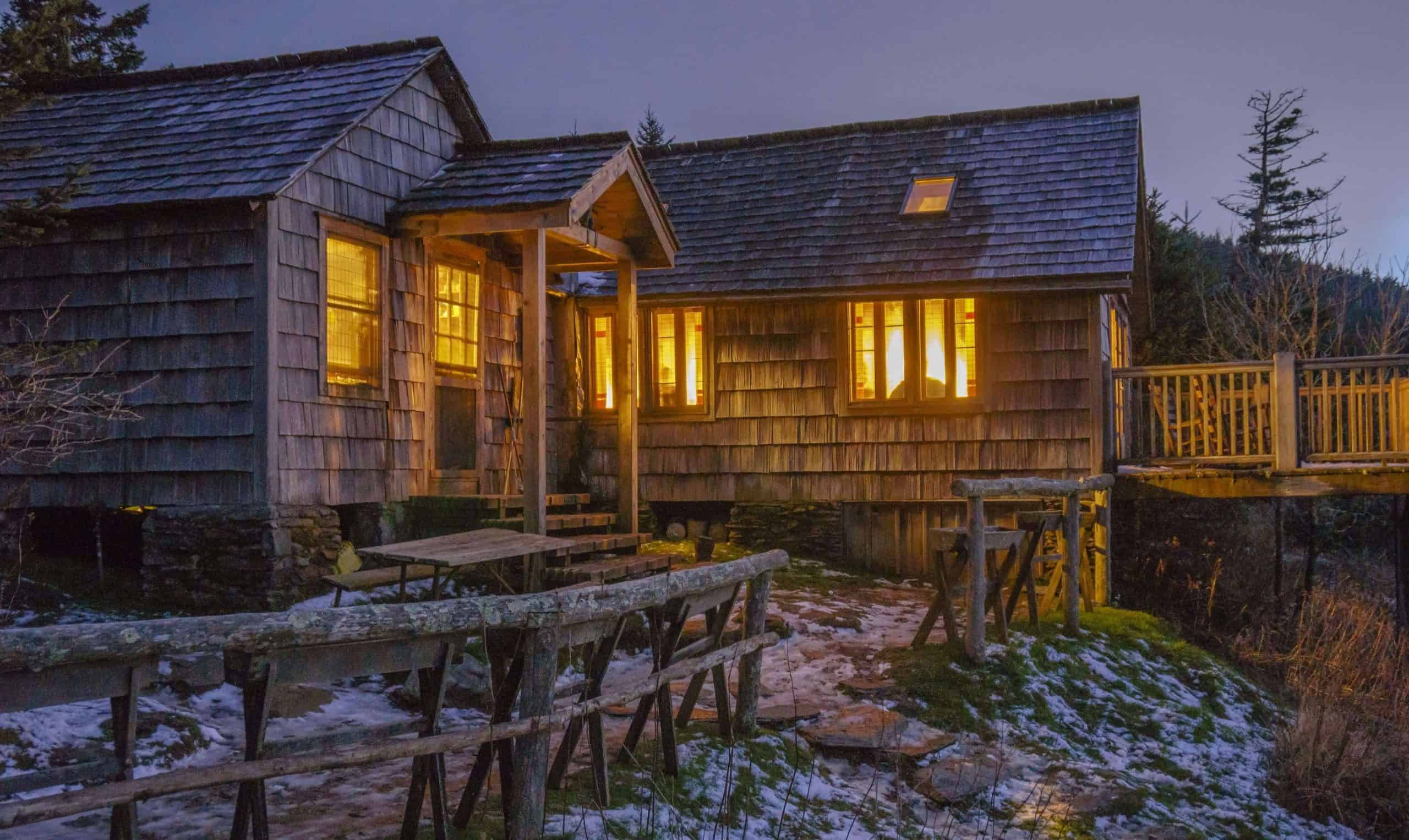 LeConte Lodge at dusk: weathered shake siding and a wood-shingle roof, warm light in the multi-pane windows, a log fence and picnic table in front, snow patches on mossy ground, a wooden walkway to the right