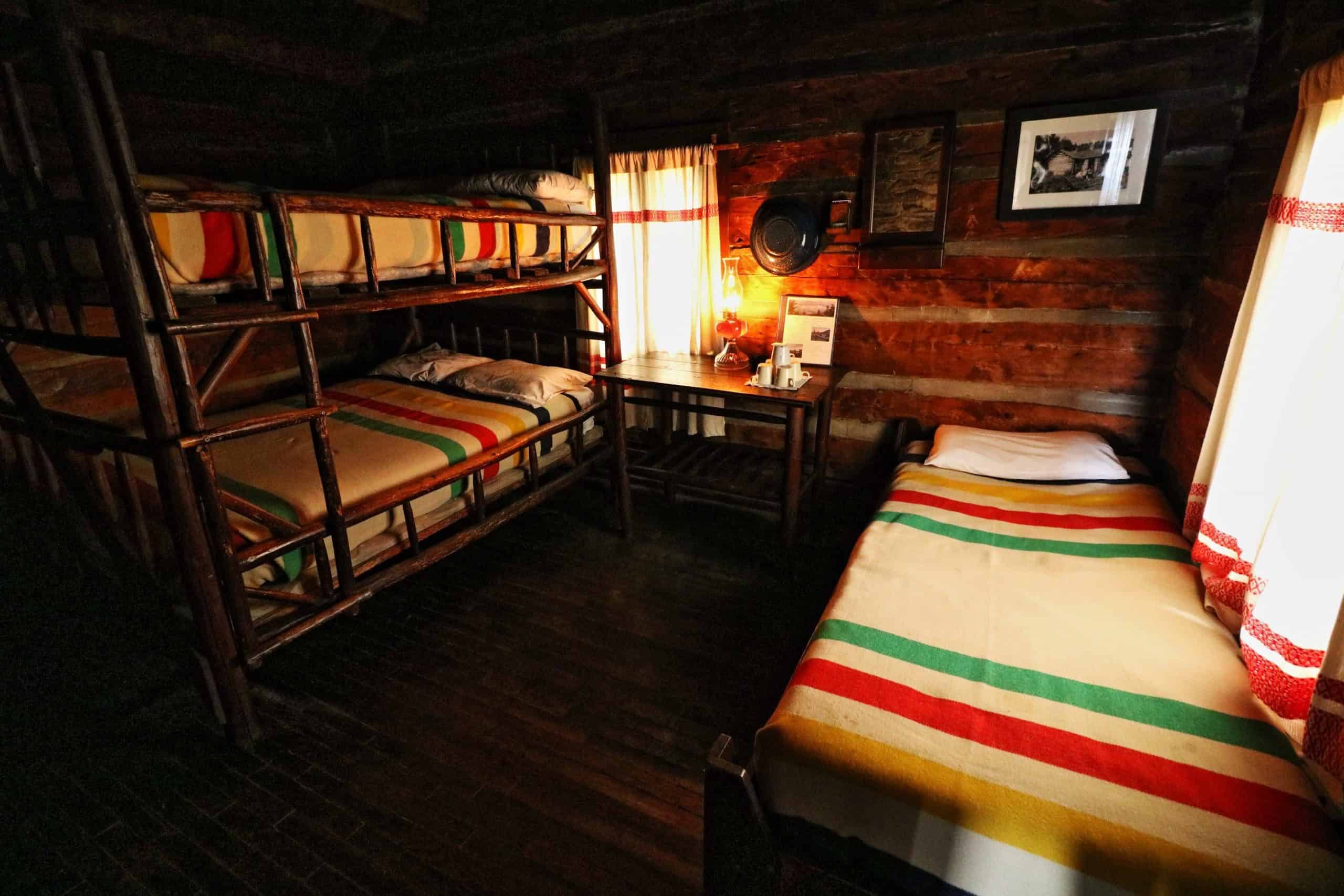 Log-cabin guest room: bunks on the left and a single bed on the right, both with Hudson Bay striped blankets, a table between them with a lit kerosene lantern, a white pitcher and mugs, chinked log walls
